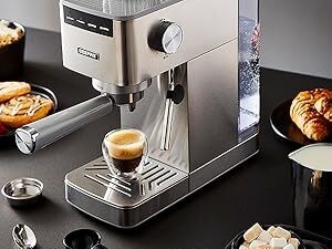 Stainless steel espresso machine on a dark counter. Features include a portafilter, steam wand, and control dial. Espresso shot and pastries visible nearby.