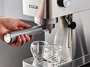 Stainless steel espresso machine in use. Hand operates portafilter while two glass cups sit on drip tray. Machine has professional appearance with control knobs visible.