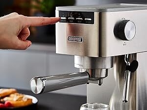 Stainless steel espresso machine with portafilter. Hand pressing button on control panel. Knob visible on side for adjustments.