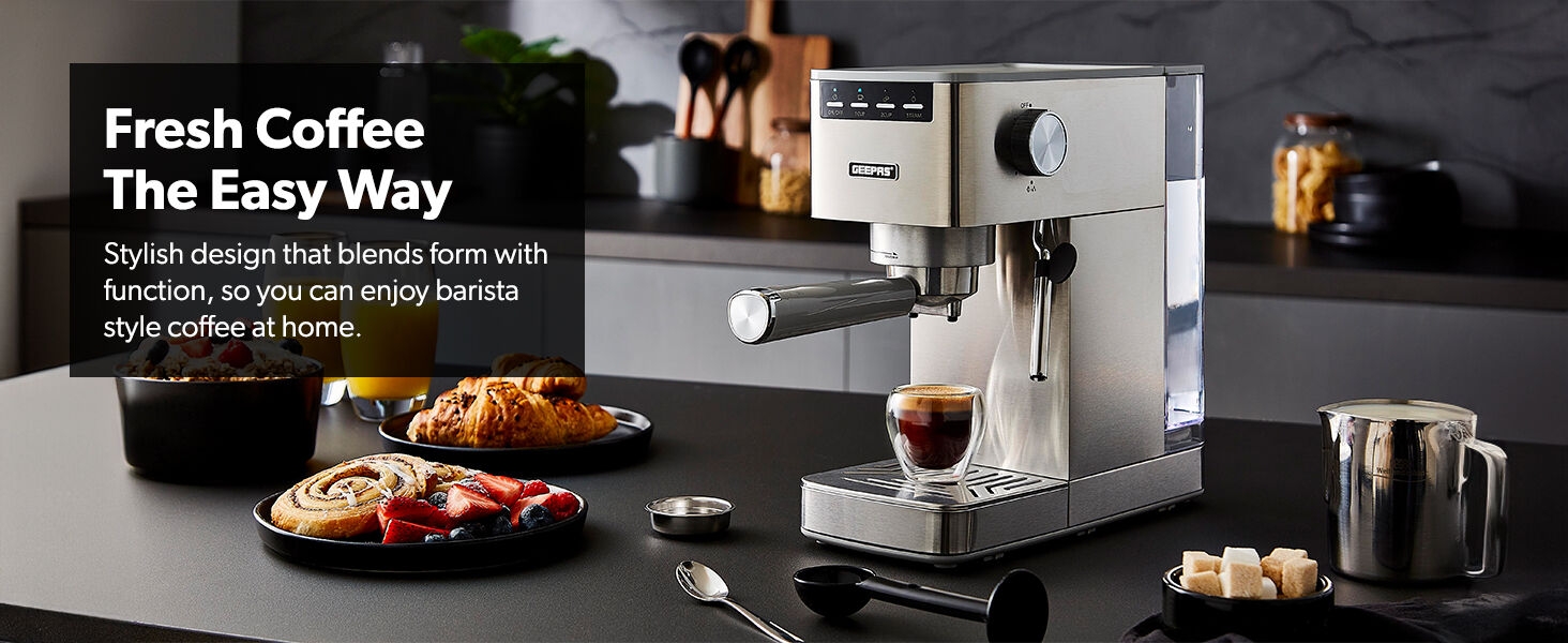 Stainless steel espresso machine with milk frother, displayed on kitchen counter with coffee, pastries, and accessories.