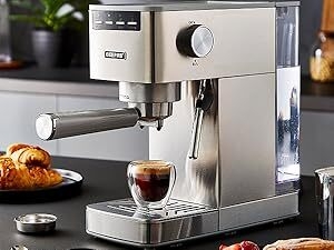 Stainless steel espresso machine on kitchen counter. Features large dial, portafilter, and steam wand. Croissants and espresso cup visible nearby.