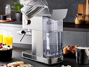 Stainless steel espresso machine with transparent water tank pouring hot water. Kitchen counter with breakfast items and orange juice visible.