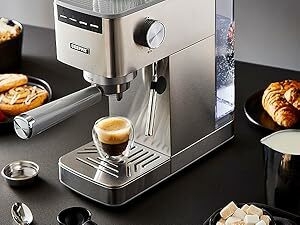 Stainless steel espresso machine on a dark counter. Features include a portafilter, steam wand, and control dial. Espresso shot and pastries visible nearby.
