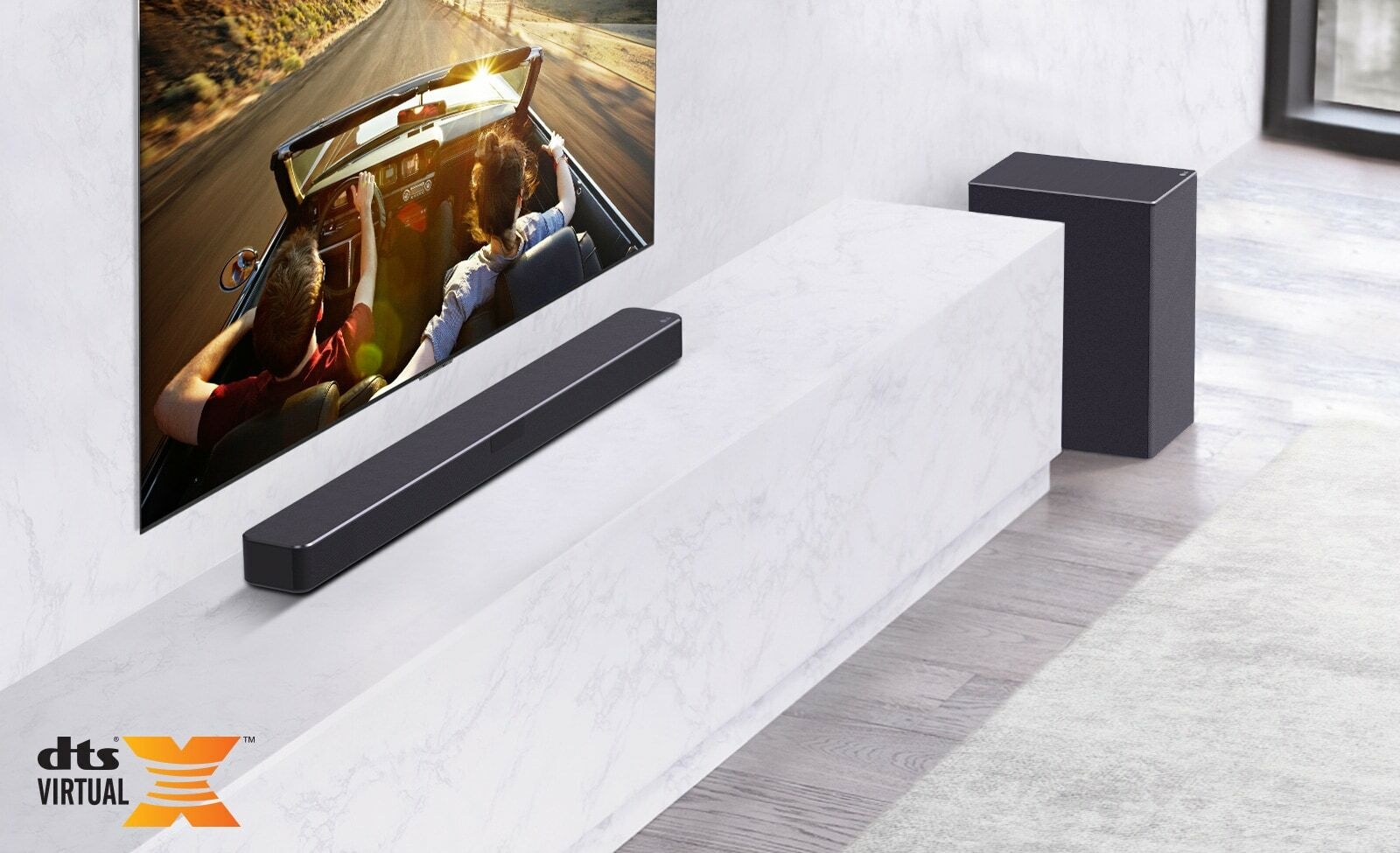 TV is on the wall, LG Soundbar is below on a white marble shelf with a sub-woofer to the right. TV shows a couple in a car.
