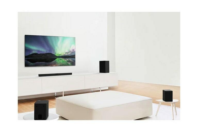 TV and sound bar in white living room with white sofa in the center. Speakers sit on both ends of the sofa.