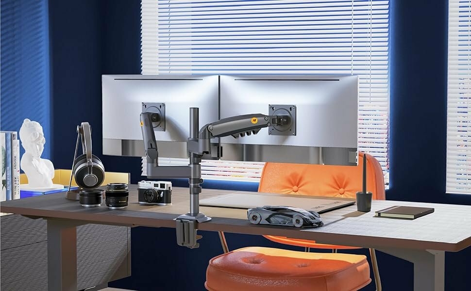 Ergonomic workspace with dual monitor setup, adjustable monitor arms, desk with orange chair, and various accessories like headphones and decorative items.