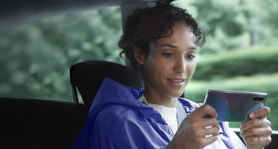 Woman sitting in a car holding Galaxy Note10 plus in landscape mode. Download manager notifications appear showing three large files being downloaded at the same time thanks to the fast data capabilities